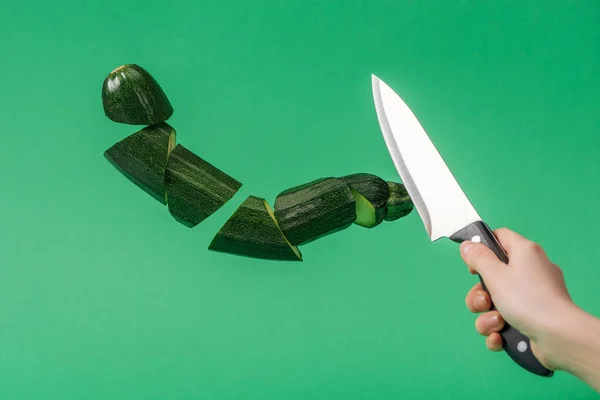 Vista cortada de mulher segurando faca com abobrinha verde fresco fatiado isolado no verde — Fotografia de Stock