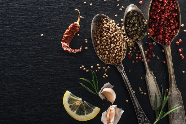 Top view of pepper and mustard in silver spoons on black textured background with dried chili pepper, lemon, herbs and garlic — Stock Photo