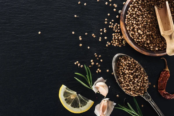 Vista superior de sementes de coentro em colher de prata e tigela de madeira em fundo preto texturizado com pimenta seca, limão, ervas e alho — Fotografia de Stock