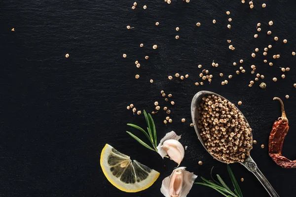 Top view of coriander seeds in silver spoon on black textured background with dried chili pepper, lemon, herbs and garlic — Stock Photo