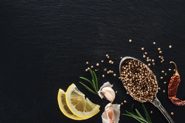 Top view of coriander seeds in silver spoon on black background with dried chili pepper, lemon, herbs and garlic — Stock Photo