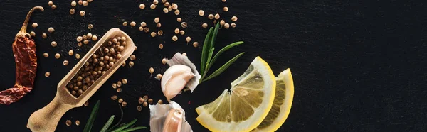 Panoramic shot of coriander seeds in wooden spatula on black textured background with dried chili pepper, lemon, herbs and garlic — Stock Photo