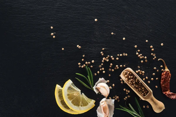 Top view of coriander seeds in wooden spatula on black textured background with dried chili pepper, lemon, herbs and garlic — Stock Photo