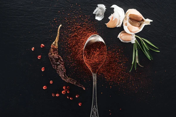Top view of red pepper powder in silver spoon on black background with dried chili peppe, herbs and garlic — Stock Photo