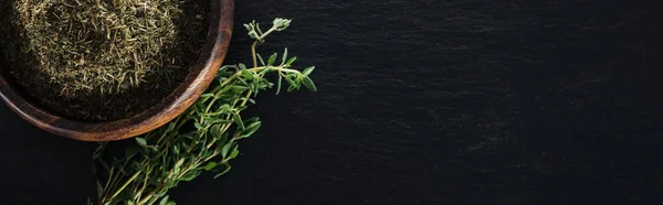 Tiro panorâmico de tomilho seco em tigela de madeira perto de erva verde no fundo preto — Fotografia de Stock