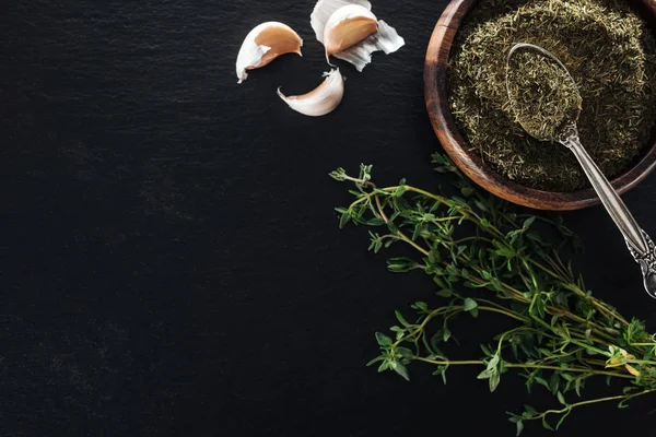 Top view of dried thyme in wooden bowl with silver spoon near green herb and garlic cloves on black background — Stock Photo