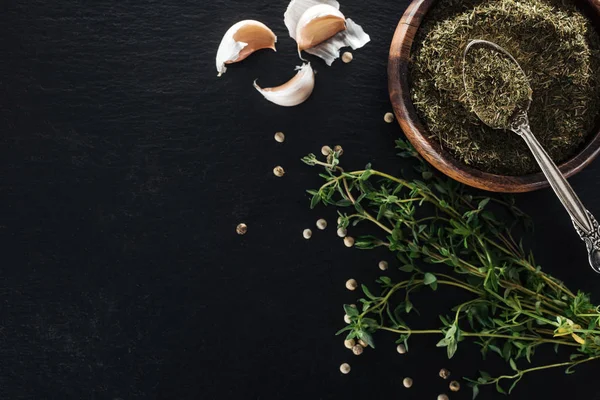 Vue du dessus du thym séché dans un bol en bois avec cuillère en argent près de l'herbe verte, poivre blanc et gousses d'ail sur fond noir — Photo de stock