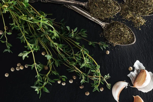 Top view of dried thyme in silver spoons near green herb, white pepper and garlic cloves on black background — Stock Photo