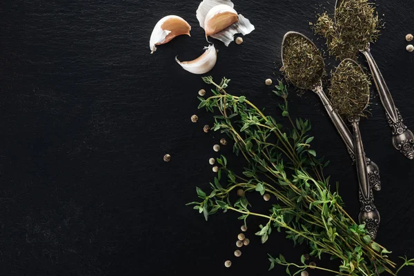 Vista dall'alto del timo essiccato in cucchiai d'argento vicino a erba verde, pepe bianco e spicchi d'aglio su sfondo nero con spazio per copiare — Foto stock