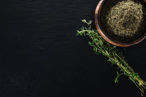 Top view of thyme spice in wooden bowl near green herb on black background — Stock Photo