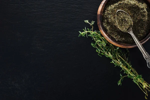 Vue du dessus du thym séché dans un bol en bois avec cuillère près de l'herbe verte sur fond noir — Photo de stock