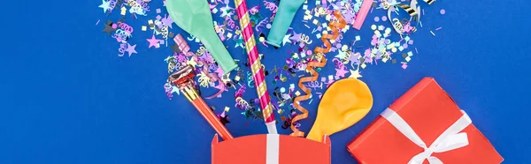 Panoramic shot of red gift box, confetti, balloons and party horns on blue background — Stock Photo