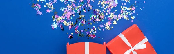 Panoramic shot of red gift box and multicolored confetti on blue background, surprise concept — Stock Photo