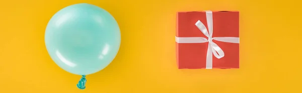 Panoramic shot of blue balloon and red gift box on yellow background — Stock Photo