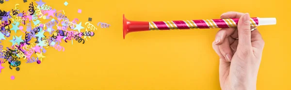 Panoramic shot of woman holding party horn near confetti on yellow background — Stock Photo