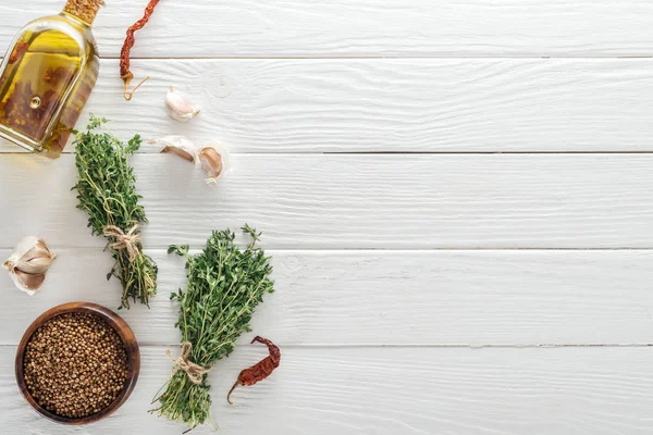 Vista dall'alto del timo verde vicino al coriandolo in ciotola, peperoncini secchi, olio in bottiglia e spicchi d'aglio sul tavolo di legno bianco — Foto stock