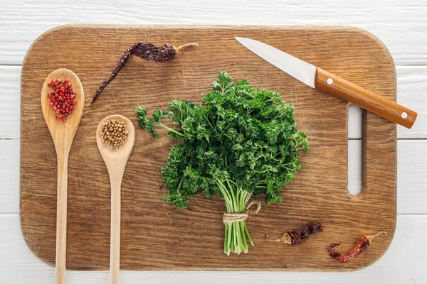 Vista superior de perejil verde, cuchillo, cucharas con cilantro y pimienta rosa, chiles secos sobre tabla de cortar de madera - foto de stock