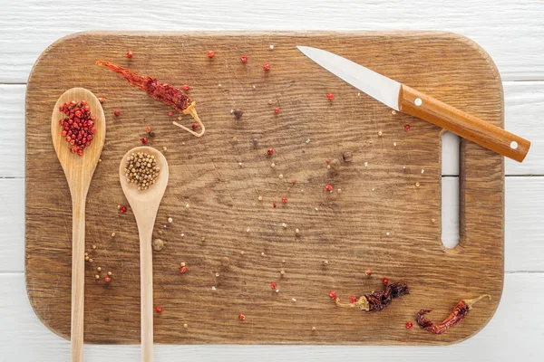 Top view of knife, spoons, scattered coriander and pink peppercorn, dried chili peppers on wooden chopping board — Stock Photo