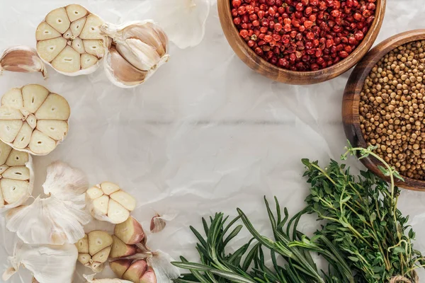 Top view of garlic cloves, pink peppercorn and coriander in bowls, rosemary and thyme on white paper background — Stock Photo