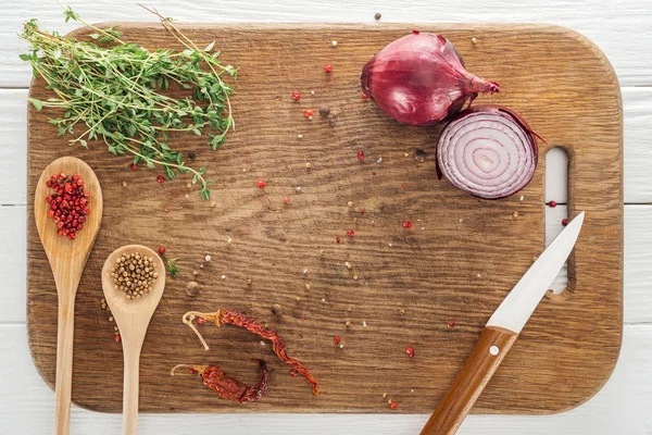 Top view of thyme, knife, spoons with coriander and pink peppercorn, dried chili peppers and red onion on wooden chopping board — Stock Photo