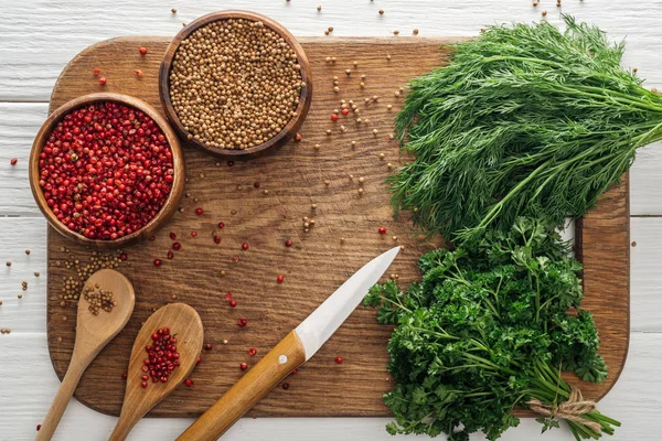 Top view of green parsley and dill, knife, spoons, coriander and pink peppercorn in bowls on wooden chopping board — Stock Photo