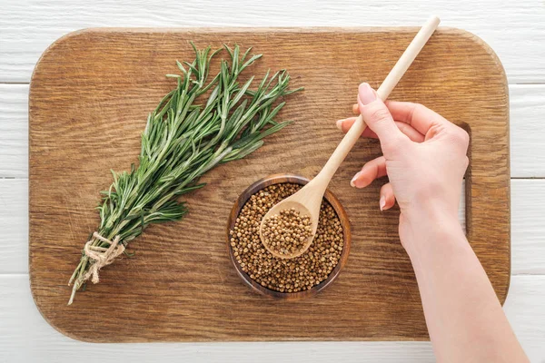 Vista ritagliata della donna che tiene cucchiaio con coriandolo vicino al rosmarino sul tagliere di legno — Foto stock