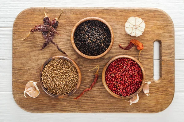 Vista superior de pimienta negra, cilantro y pimienta rosa en cuencos sobre tabla de cortar de madera - foto de stock