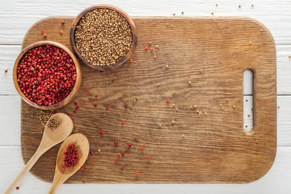 Top view of coriander and pink peppercorn in bowls and spoons on wooden chopping board — Stock Photo