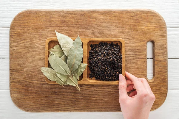 Vue recadrée d'une femme touchant des bols en bois avec des feuilles de laurier et du poivre noir sur une planche à découper — Photo de stock