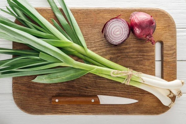 Top view of green leek and red onion on wooden chopping board with knife — Stock Photo
