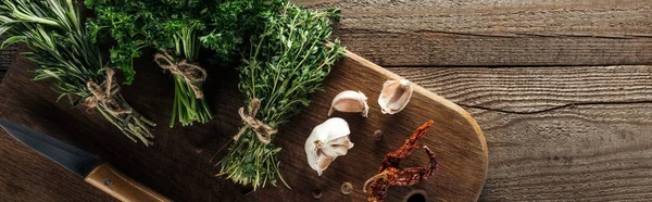 Panoramic shot of green thyme, parsley, rosemary on wooden chopping board near garlic cloves, knife, dried chili pepper and black pepper on wooden brown table — Stock Photo
