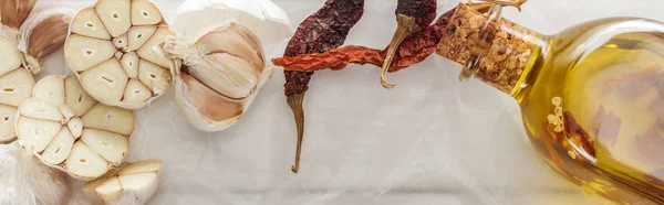 Panoramic shot of garlic cloves, dried chili peppers and bottle with oil on white paper background — Stock Photo