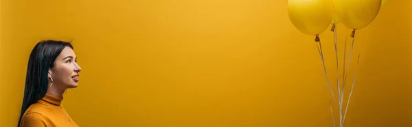 Улыбающаяся девушка держит праздничные минималистичные воздушные шары на желтом фоне, панорамный снимок — стоковое фото
