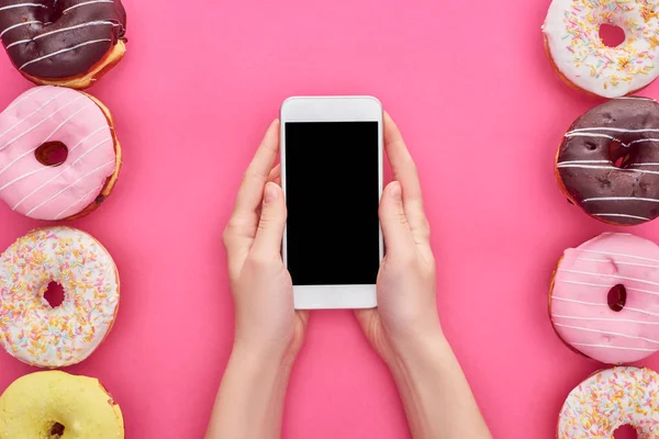 Vista ritagliata di donna che tiene smartphone con schermo bianco vicino gustose ciambelle smaltate su sfondo rosa brillante — Foto stock