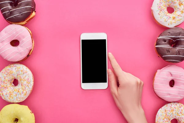 Обрезанный вид женщины, указывающей пальцем на смартфон на розовом фоне с вкусными пончиками — стоковое фото