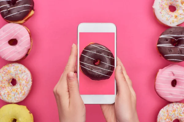 Vue recadrée de femme tenant smartphone avec image beignet sur fond rose près de beignets savoureux — Photo de stock
