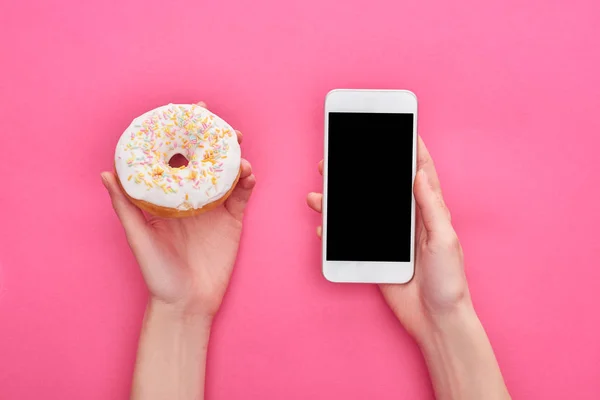 Vista parcial de la mujer sosteniendo smartphone con pantalla en blanco cerca de sabrosa rosquilla acristalada con aspersiones sobre fondo rosa brillante - foto de stock