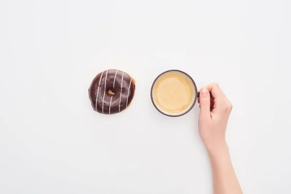 Vista parcial de la mujer sosteniendo la taza con café cerca de sabrosa rosquilla de chocolate esmaltado sobre fondo blanco - foto de stock