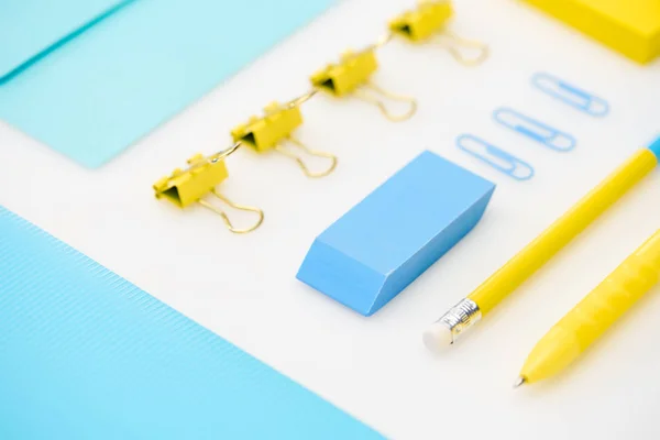 Couche plate de gomme bleue, trombones, dossier, enveloppe, stylo jaune, crayon, autocollants en fond blanc — Photo de stock