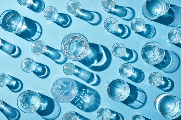 Draufsicht auf Gläser mit klarem Wasser und Schatten auf blauer Oberfläche — Stockfoto