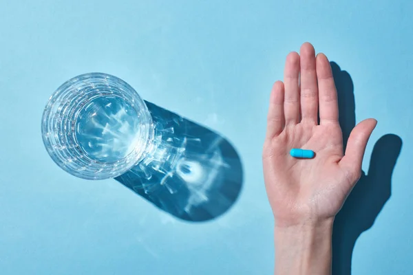 Vista recortada de la mujer sosteniendo la píldora azul en la palma cerca del vidrio con agua sobre fondo azul - foto de stock