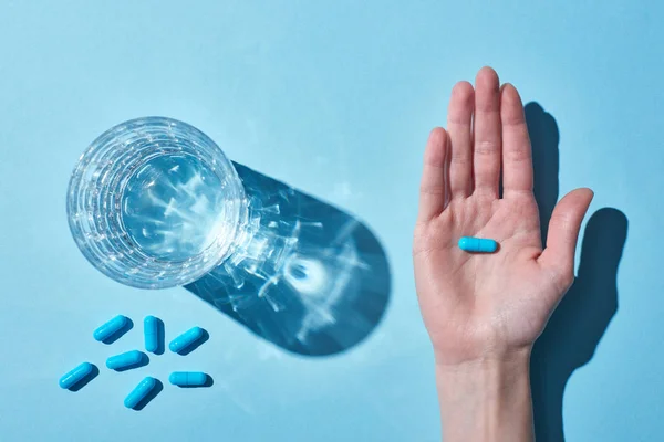 Partial view of woman holding blue pill on palm near glass with water on blue background — Stock Photo