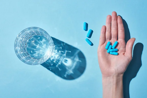 Vista ritagliata della donna che tiene pillole blu sul palmo vicino al vetro con acqua su sfondo blu — Foto stock