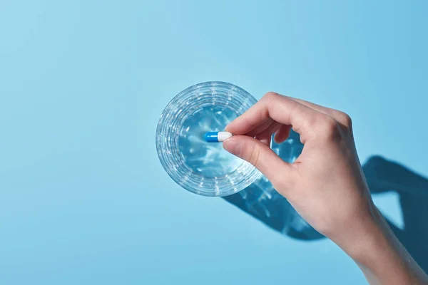 Vue recadrée de la femme mettant pilule en verre avec de l'eau sur fond bleu — Photo de stock