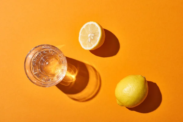 Vista dall'alto di vetro con acqua vicino limoni gialli su sfondo arancione — Foto stock