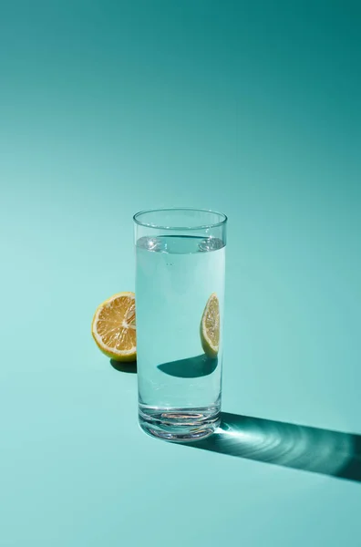 Transparent glass with fresh water and yellow lemon half on turquoise background — Stock Photo