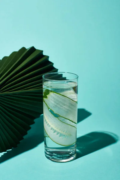 Transparent glass with fresh water and cucumber slices near green leaf on turquoise background — Stock Photo