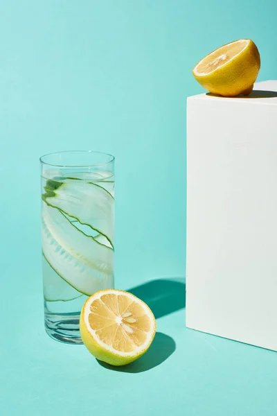 Transparent glass with pure water and cucumber slices near lemons on turquoise background — Stock Photo