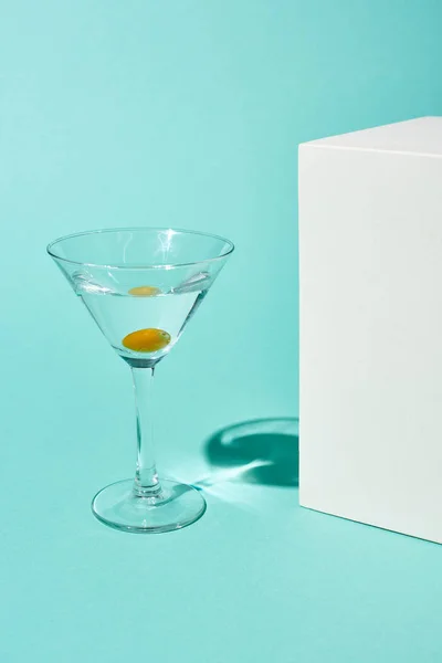 Transparent glass with cocktail and olive near white cube on turquoise background — Stock Photo