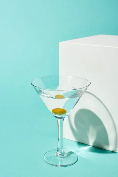Transparent glass with cocktail and olive near white cube on turquoise background — Stock Photo